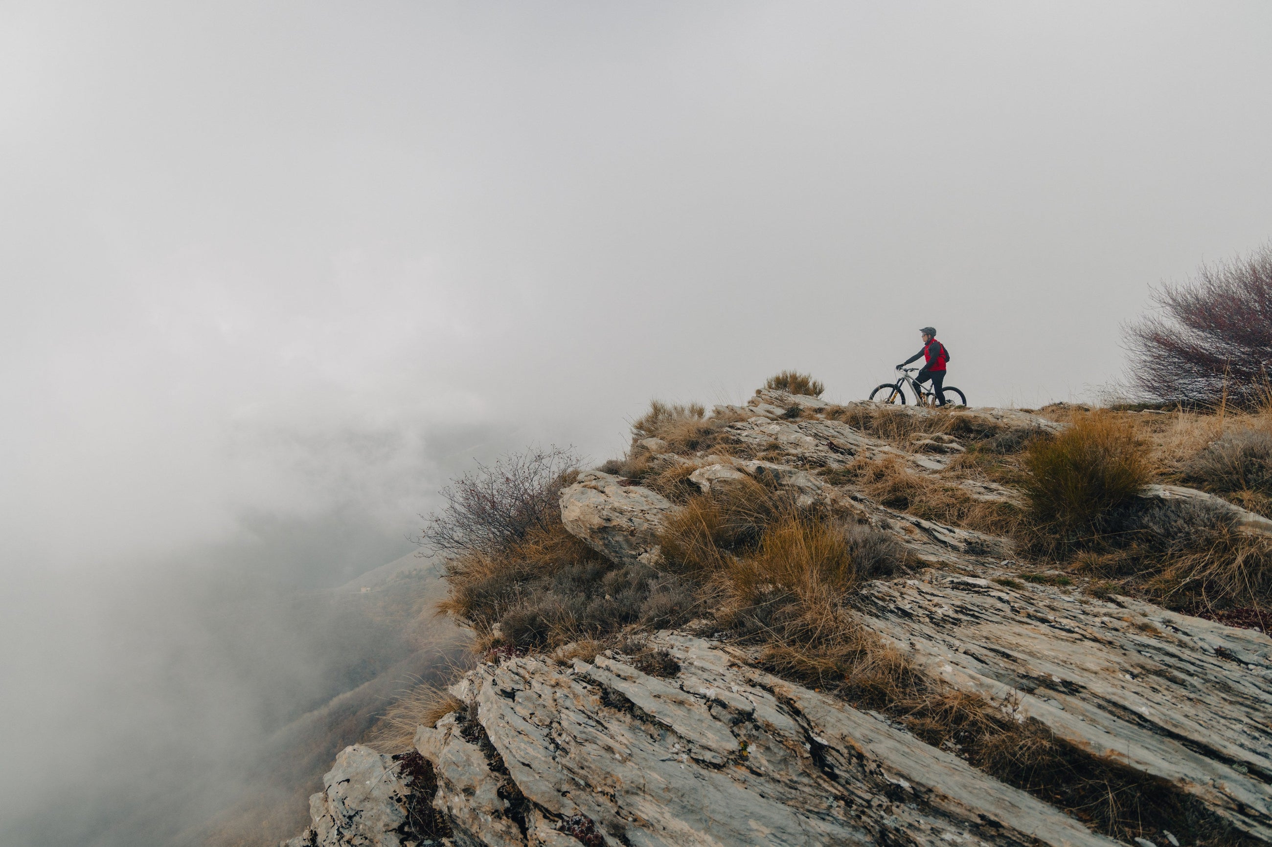 A rider standing over a mountain bike on a rocky ridge above the clouds.