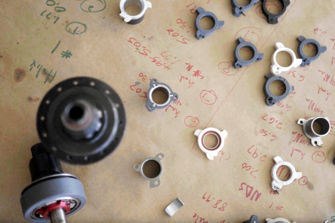 Dozens of 3D printed pusher prototypes laid out on a workbench covered in handwritten measurements, with a Sidekick hub and axle in the foreground.