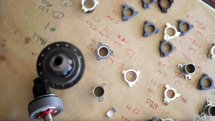 Dozens of 3D printed pusher prototypes laid out on a workbench covered in handwritten measurements, with a Sidekick hub and axle in the foreground.