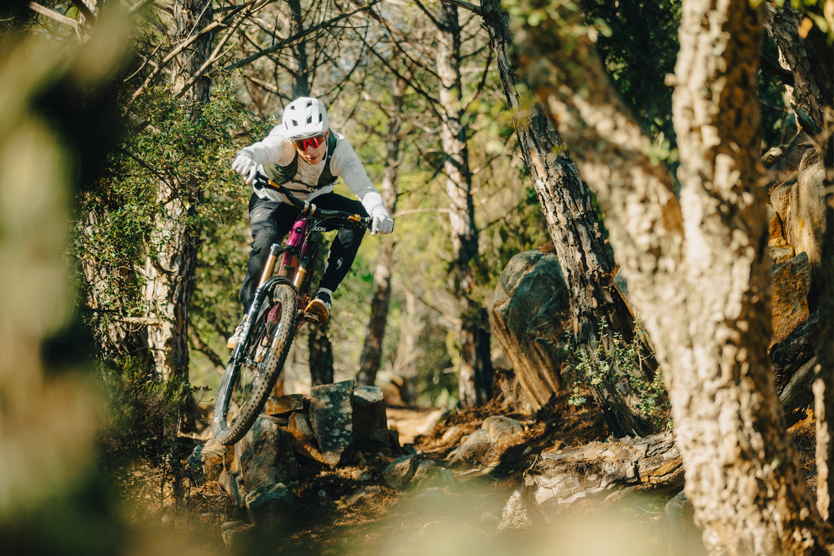 Rider lifting the front wheel over roots on a forest trail, all-mountain terrain.