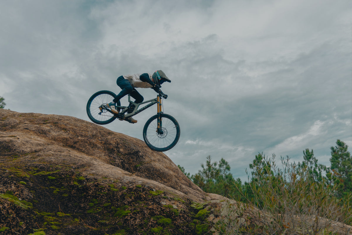 Downhill rider launching off a granite slab under a grey sky.